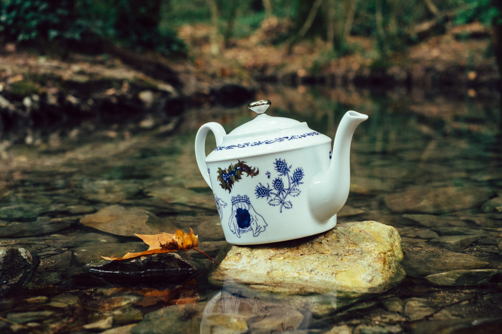 Blue teapot from “Enchanteresse”, a collection of painted porcelains by messalyn, standing on a rock in a small pool in the forest